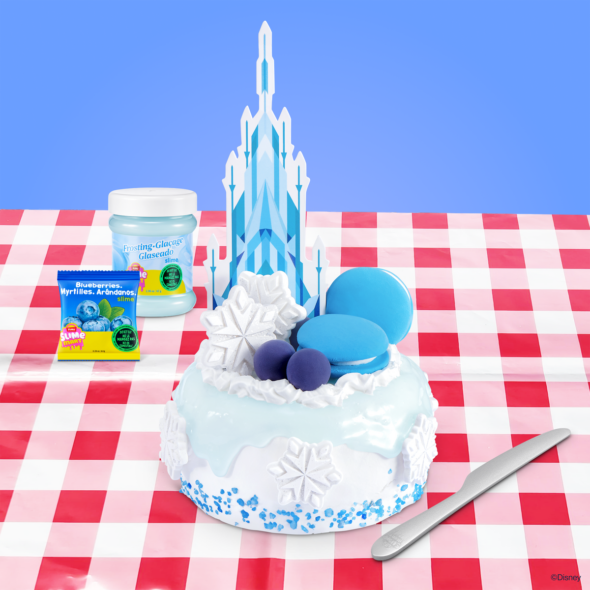 Icy blue slime cake with white frosting, frosting tools, and slime jars on check pattern mat