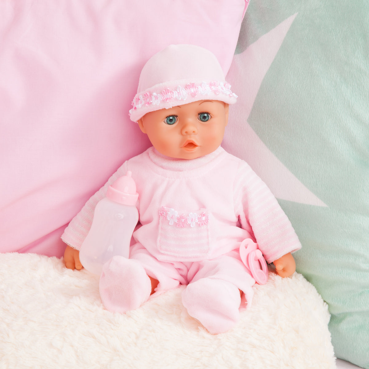 Pink baby doll with frilly hat, long-sleeve outfit, and pacifier, seated on blanket.