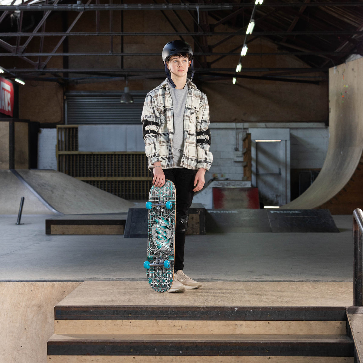 Person holding a complete skateboard with dark graphic deck and teal wheels