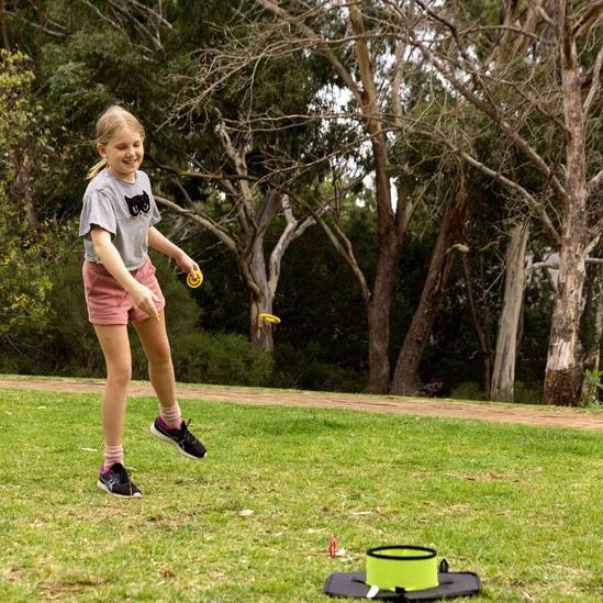 Child outdoors throwing beanbags toward green cube and yellow target with poles